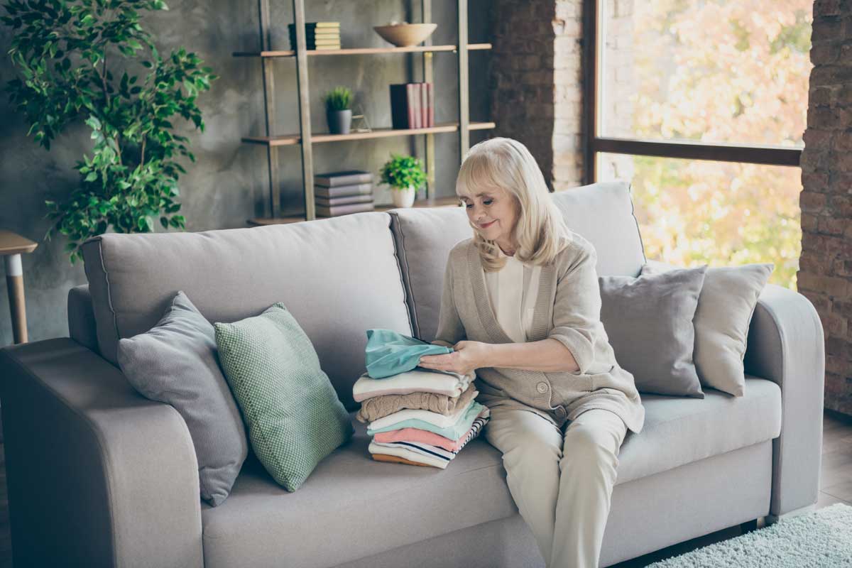 Portrait of her she nice attractive kind hardworking gray-haired granny sitting on divan folding belongings things clothing duty at industrial brick loft modern style interior house apartment